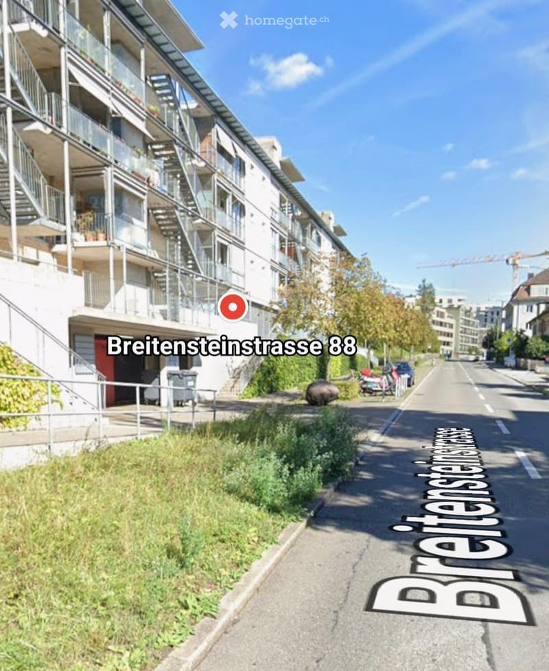 A street view of Breitensteinstrasse 88, showing a multi-story building with balconies and stairs on the left side, and a grassy area on the right side. There are two cars parked on the side of the road, and a crane is visible in the distance.