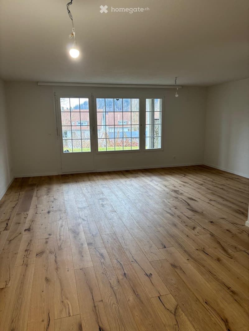 A spacious empty room with wooden floors, glass sliding doors, three windows, a white ceiling, and a ceiling light.