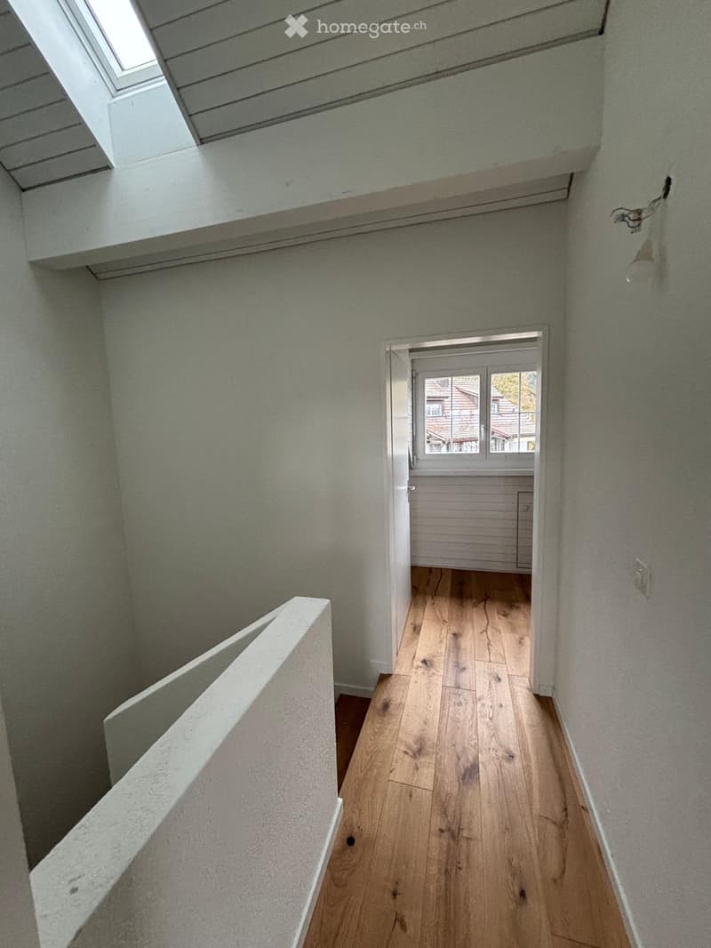 White walls, wooden floor, small hallway, light fixture on wall, open doorway, skylight, open room