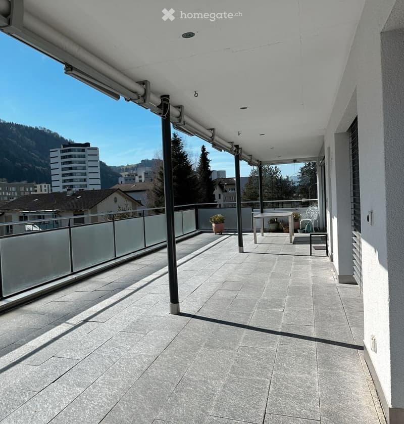 Balcony with tiled floor, metal railing, potted plants, table, chairs, and a view of the city and mountains