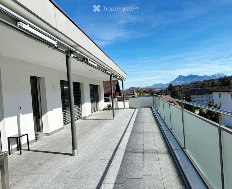 Balcony with glass railings, tiled floor, metal poles, small table, trash bin, distant mountains