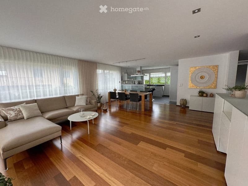 Living room with hardwood floors, large windows, white curtains, a couch, a coffee table, a dining table with chairs, a kitchen, and a decorative wall art.