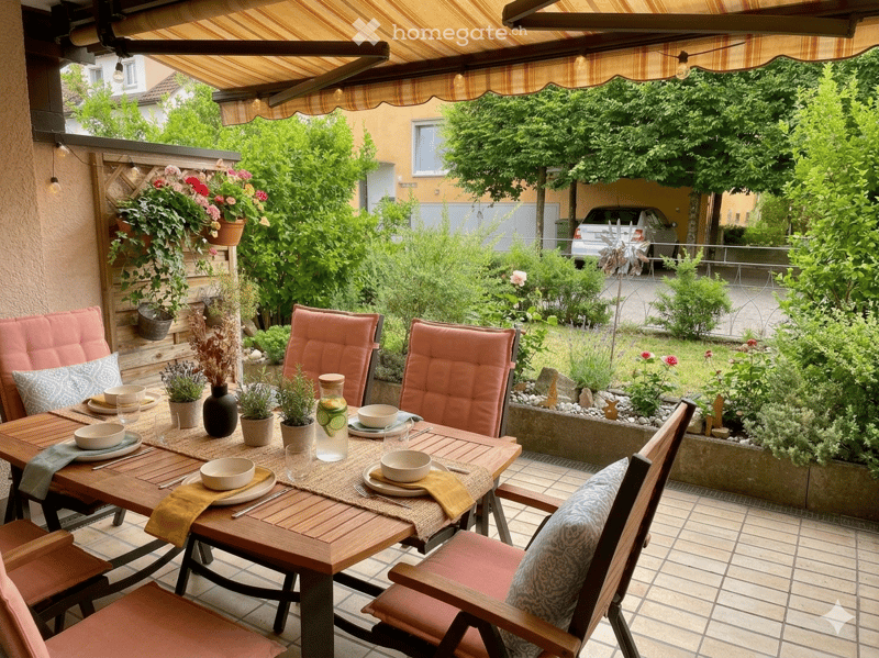 Wooden table with chairs, covered by an awning, flowers, plants, and a view of a garden and a car
