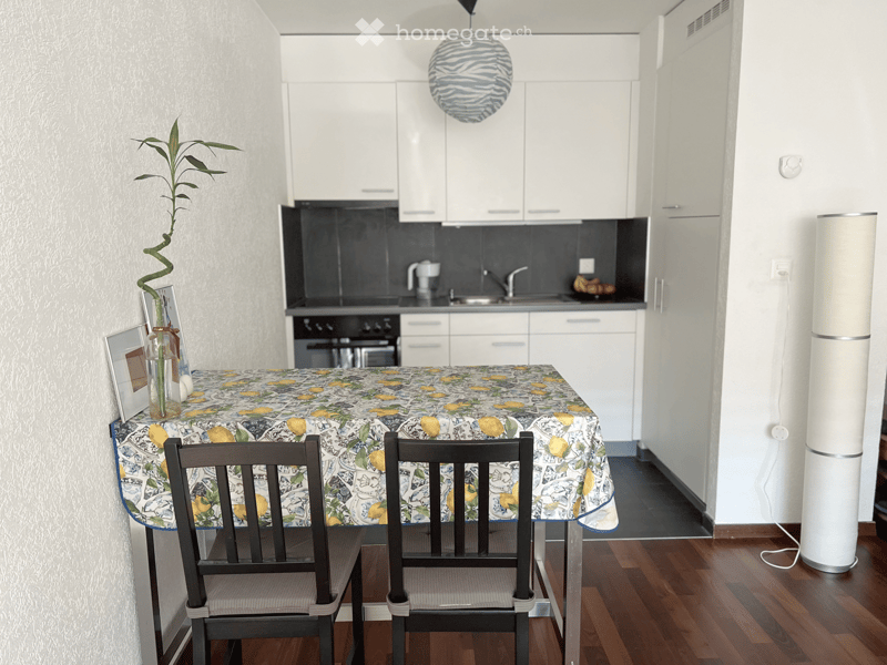 Dining area with a table, chairs, and a plant. Kitchen with white cabinets, black countertops, and a sink.