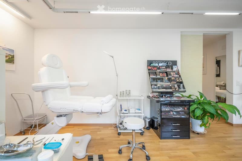A room with a white chair, a desk, a stool, a lamp, a cabinet, a trash bin, a potted plant, and a picture frame on the wall.