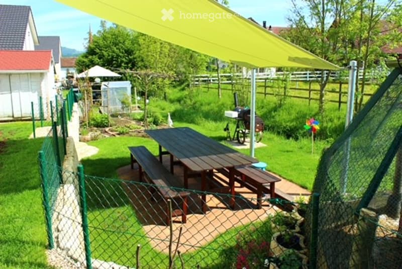 Wooden picnic table with benches, yellow canopy, green fence, plants, and a windmill