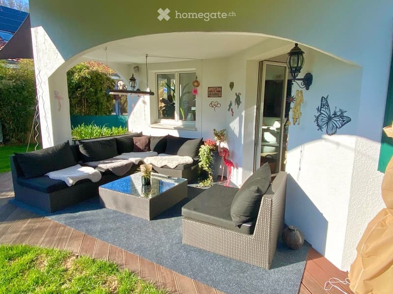 Outdoor seating area with a sofa, coffee table, and a rug, surrounded by plants and a wall with decorations