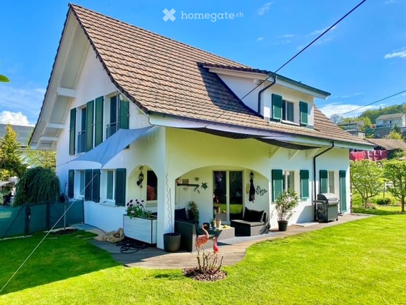 2 story house, white exterior, brown roof, green shutters, large front yard, patio with seating, garden, awning