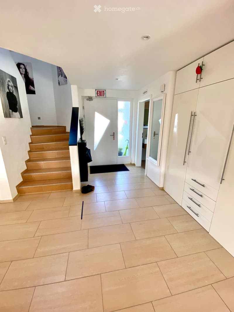 Entrance hall with wooden stairs, tiled floor, white walls, white cabinets, exit sign, glass door, black mat, picture frames, and a handbag