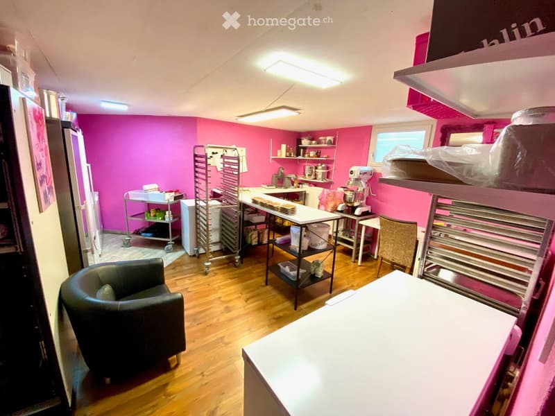 A room with pink walls, wooden floor, a black chair, a white table, shelves with various items, a mixer, and a window.