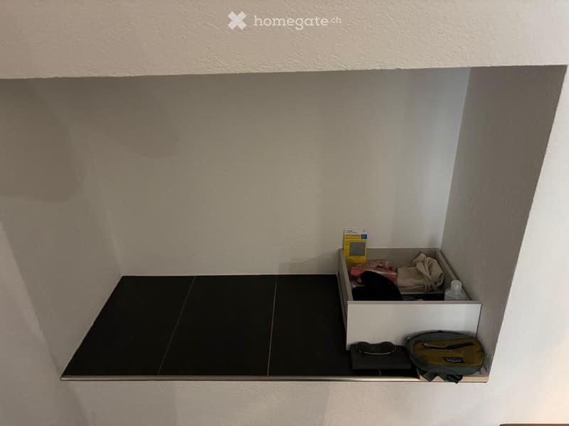 A corner shelf with a black tiled surface and a white storage box on it, containing various items such as a yellow box, a bottle, and a bag.