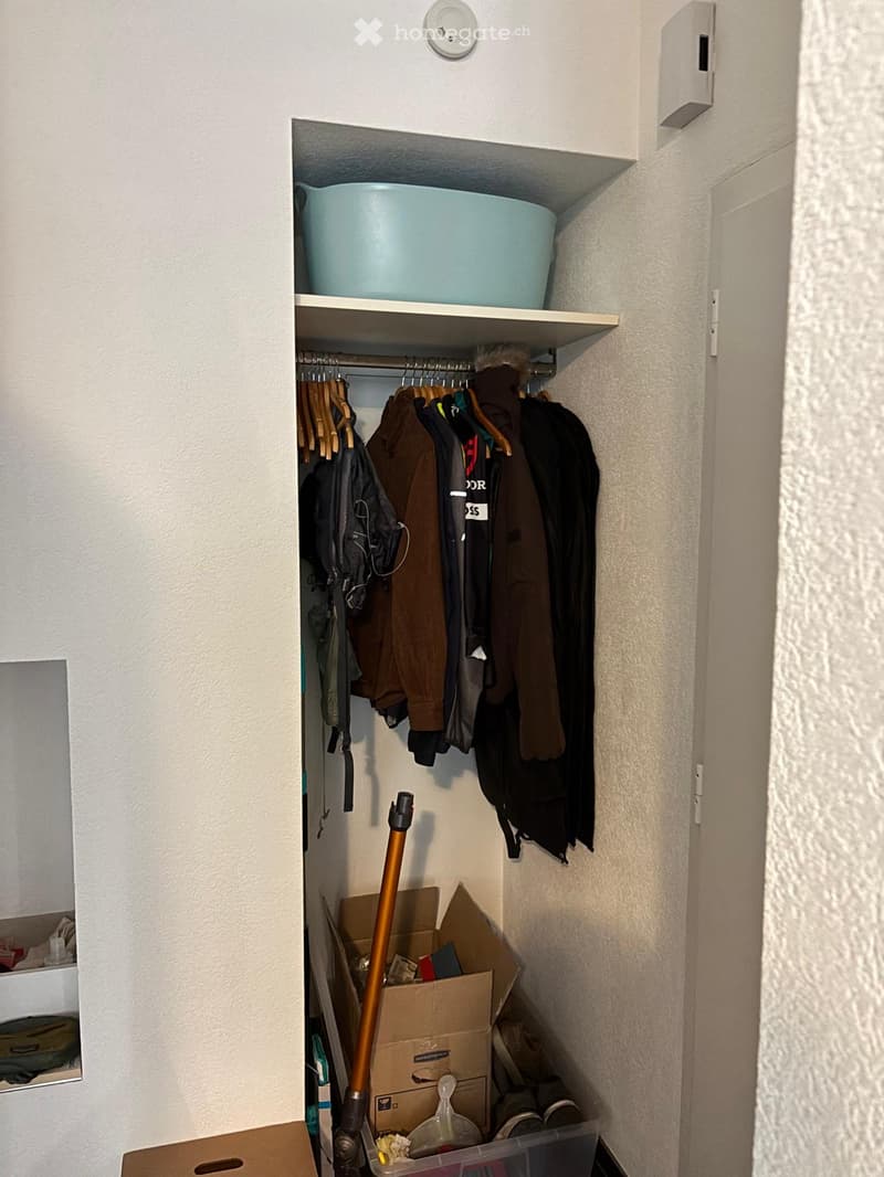 Closet with clothes hanging, storage box, and a blue bucket on the top shelf