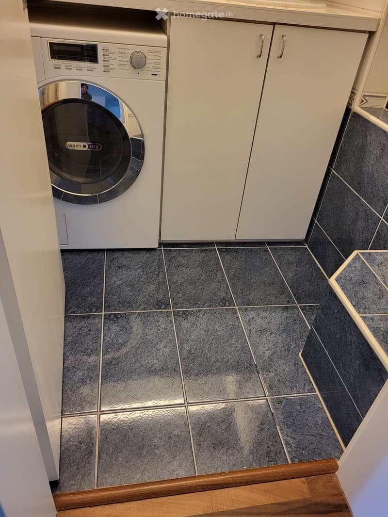A small bathroom with a white washing machine, white cabinets, and grey tiled floor.