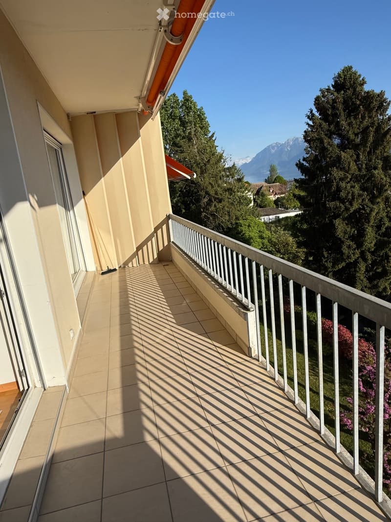 A balcony with a tiled floor and a metal railing, offering a view of a garden and mountains in the distance.