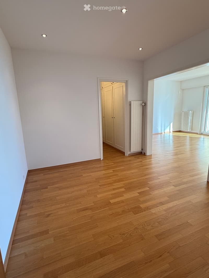An empty room with white walls, wooden floors, and recessed ceiling lights. There is a white wardrobe in the corner and a white radiator on the wall.