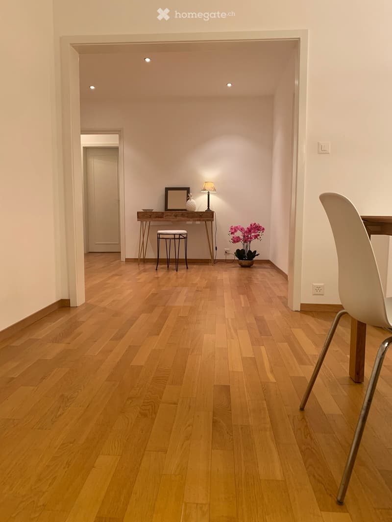 Empty room with hardwood floors, white walls, desk with lamp, stool, pink flowers in pot, white chair, power outlet