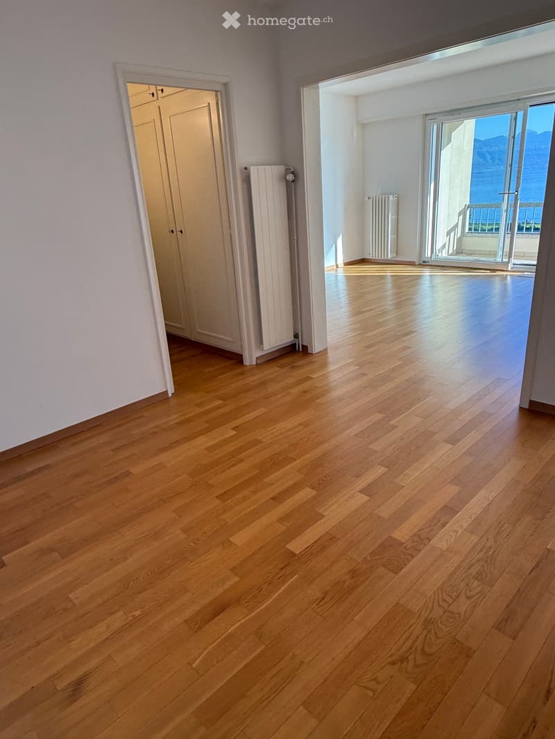 Empty room with hardwood floors, white walls, and sliding glass doors leading to a balcony with a view of the sea and mountains.