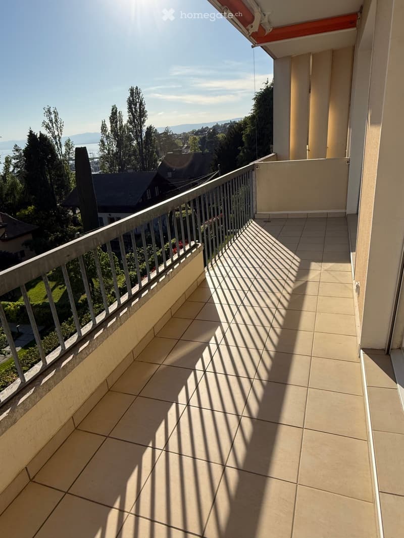 A balcony with a tiled floor and a metal railing, offering a view of trees and a distant house.