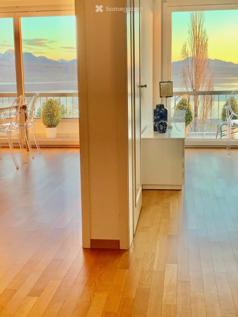 Spacious room with wooden floor, large glass windows with balcony overlooking a scenic view of mountains and water.