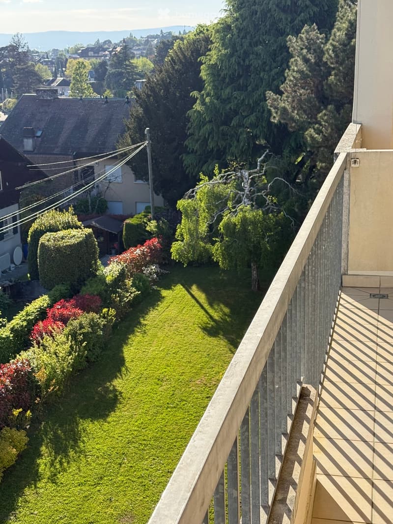 A balcony with a metal railing and a tiled floor, overlooking a well-maintained garden with a variety of plants and trees.