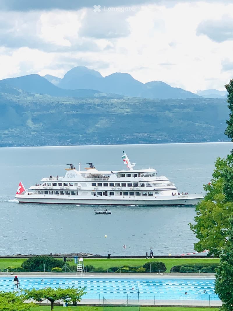 A view of a ferry boat sailing on a lake, mountains in the background, a swimming pool nearby.
