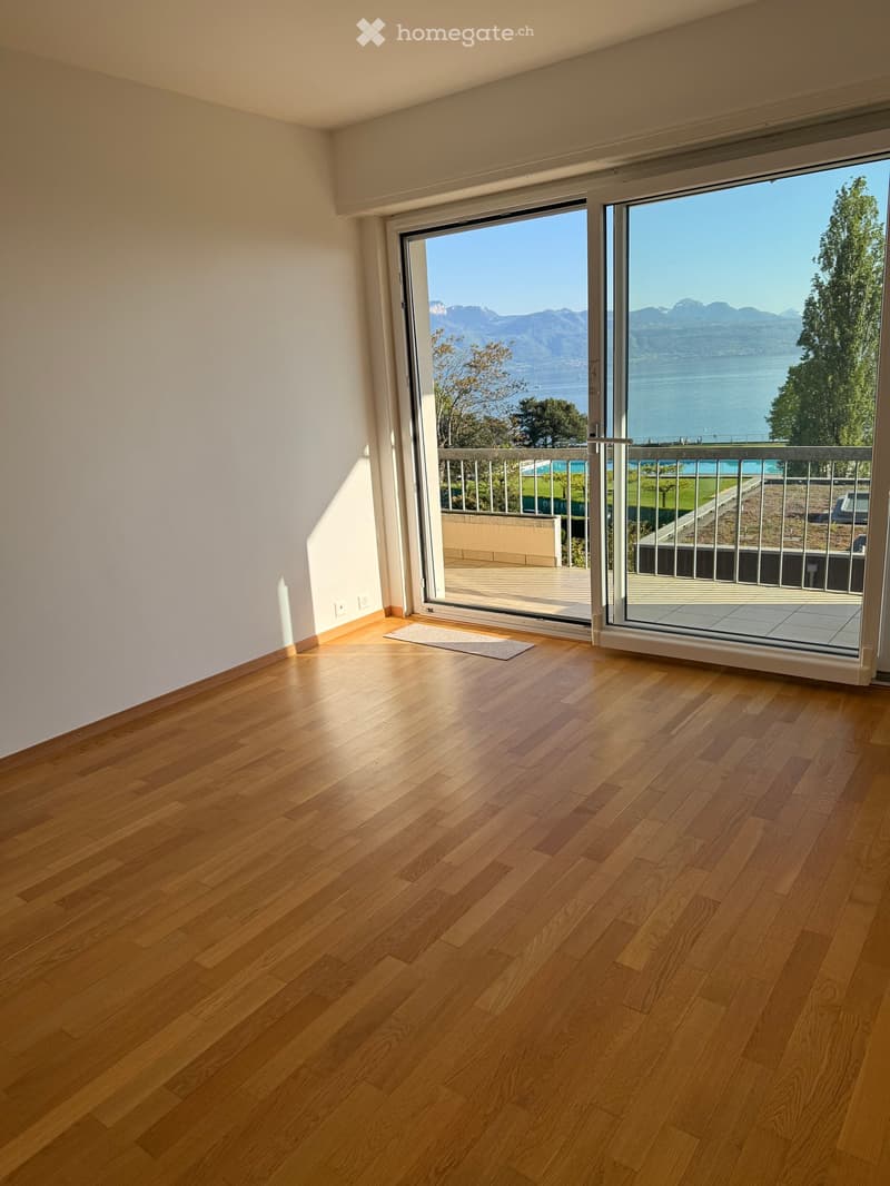 Empty room with wooden floors, white walls, and large sliding glass doors leading to a balcony with a view of a lake and mountains.