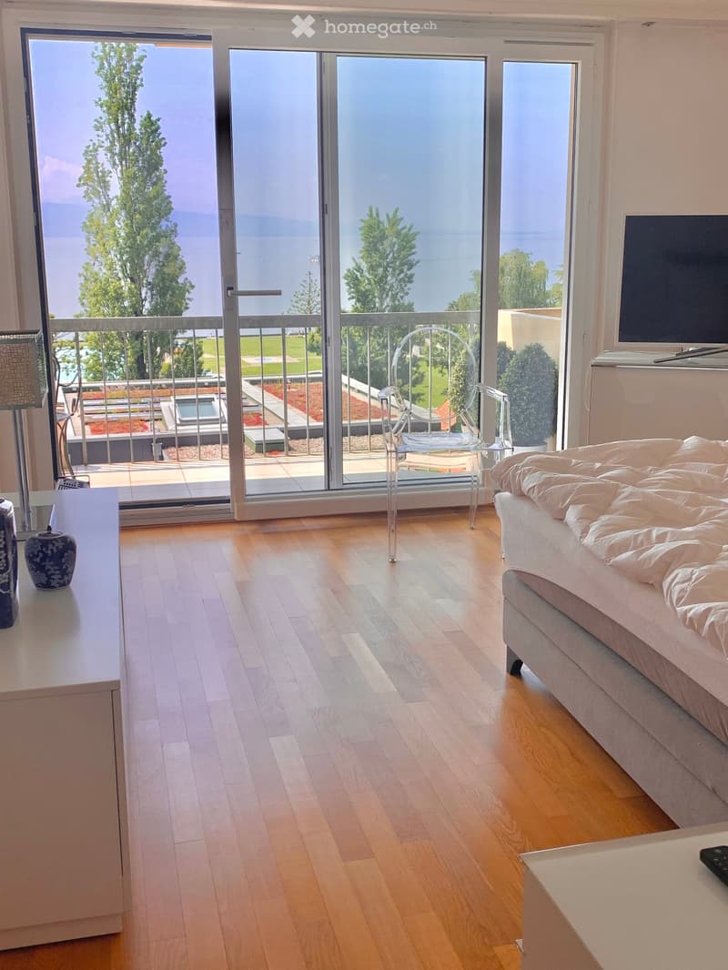 spacious room, large windows, balcony with railing, wooden floor, white walls, bed, transparent chair, television, table with decorative items