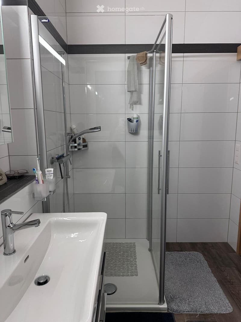 modern bathroom with white tiles, glass shower enclosure, white sink, silver faucet, towel rack, and a rug