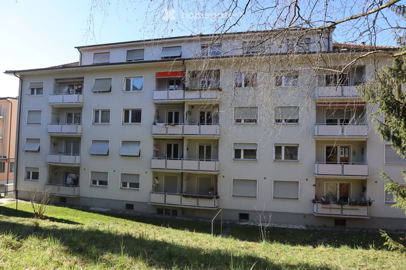 5 story apartment building, white exterior, multiple balconies, windows with shutters, small garden in front