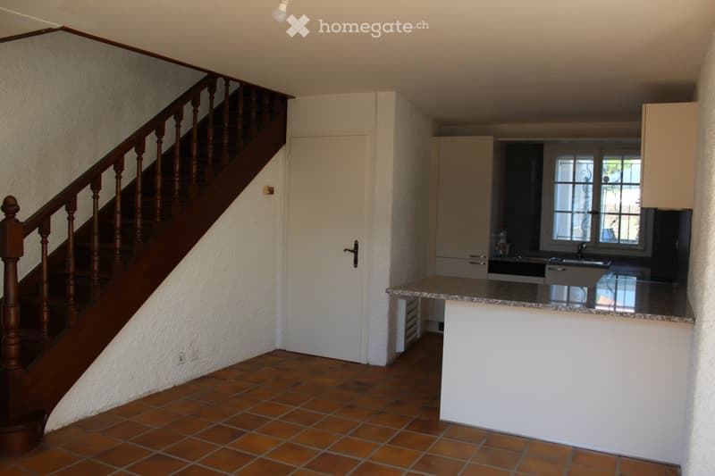 Empty room with wooden stairs, tiled floor, white walls, white door, kitchen area with countertop, cabinets, and sink.