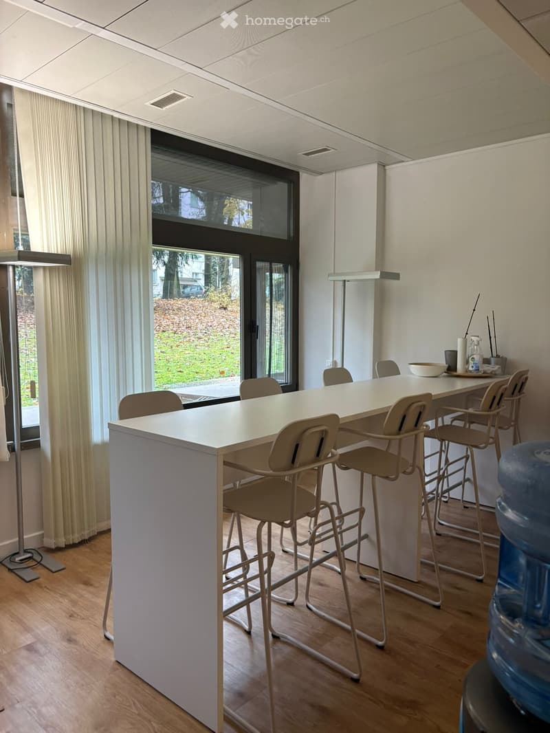 A long white table with chairs, a bowl, and a bottle on it. A lamp is on the left side, and a water dispenser is on the right side. A window with white curtains is on the left side, and a glass door is on the right side. A grassy field is outside the wind