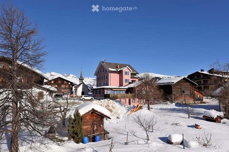 Snowy village with wooden houses, pink house in the center, mountains in the background