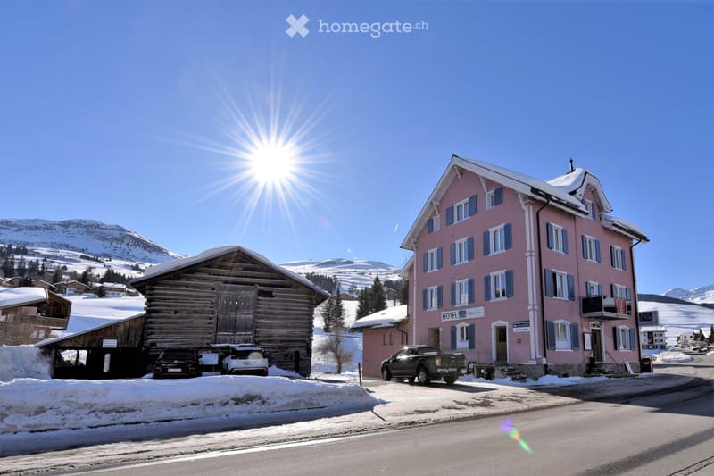 Two buildings, one pink and one wooden, with cars parked in front, snowy mountains in the background