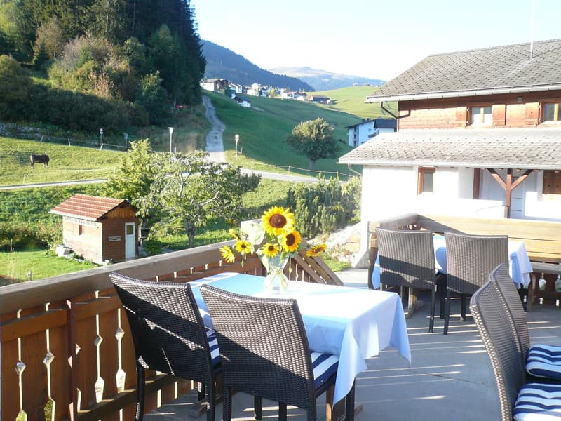 Wooden chairs and tables on a balcony with a view of a green field and mountains