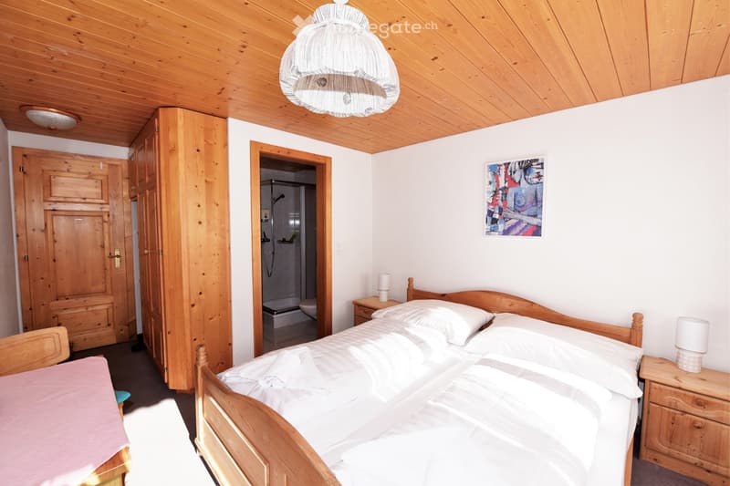Bedroom with wooden bed, wooden wardrobe, wooden nightstands, white linen, wooden ceiling, white walls, lamp, picture frame, wooden door, and a small bathroom with a shower.