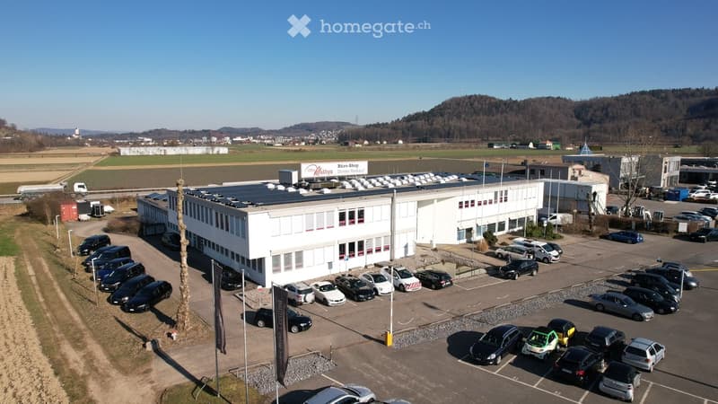 White building, solar panels on the roof, parking lot with many cars, mountains in the background