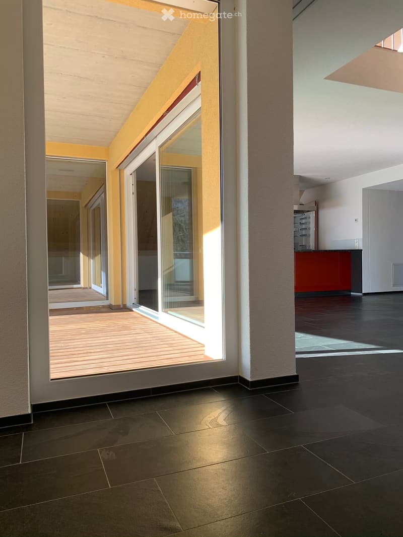 Open space with large glass doors leading to a wooden deck, modern interior design, black tiled floor, yellow walls, glass walls, and a glimpse of a kitchen area.