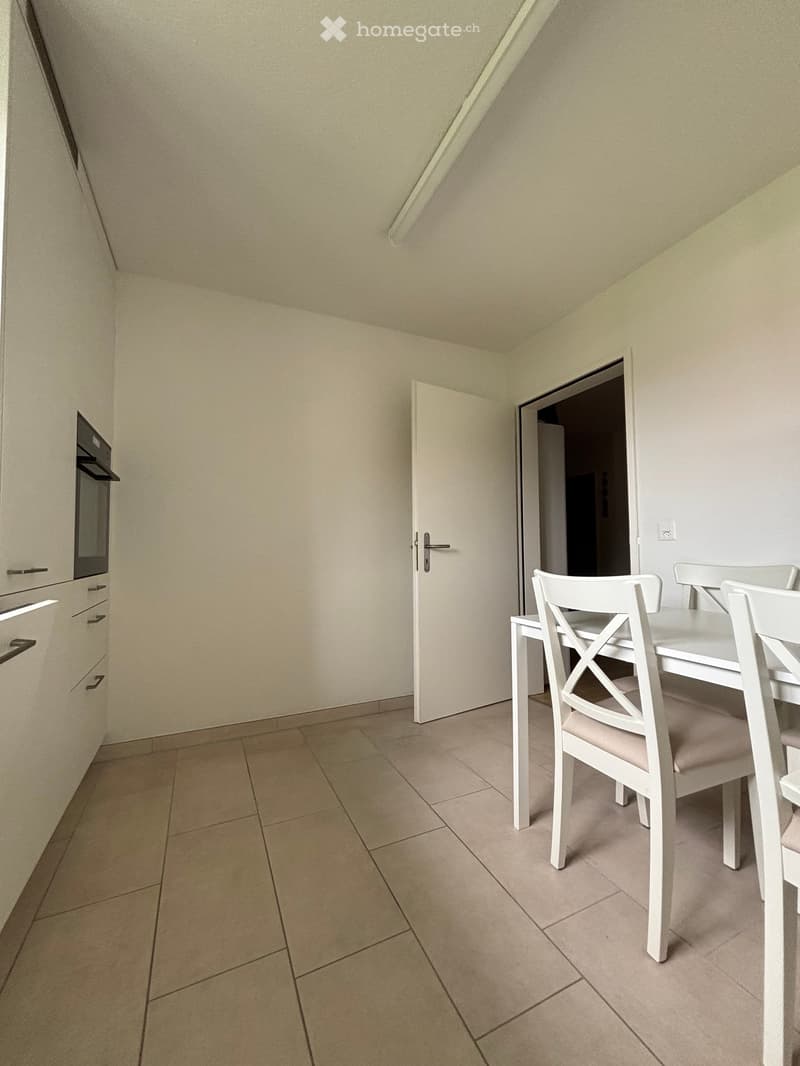 Empty room with white walls, tiled floor, white dining table and chairs, white kitchen cabinets, and an oven.