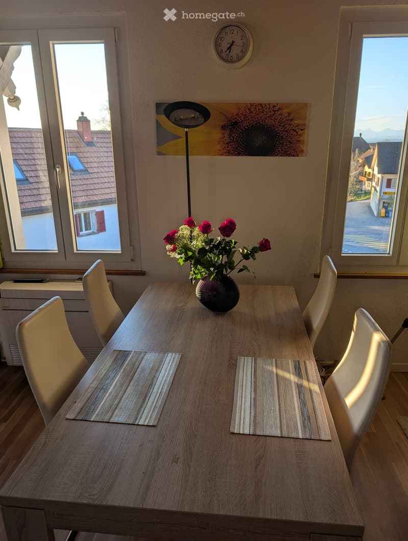 Dining room with wooden table, white chairs, flower vase, clock, and windows