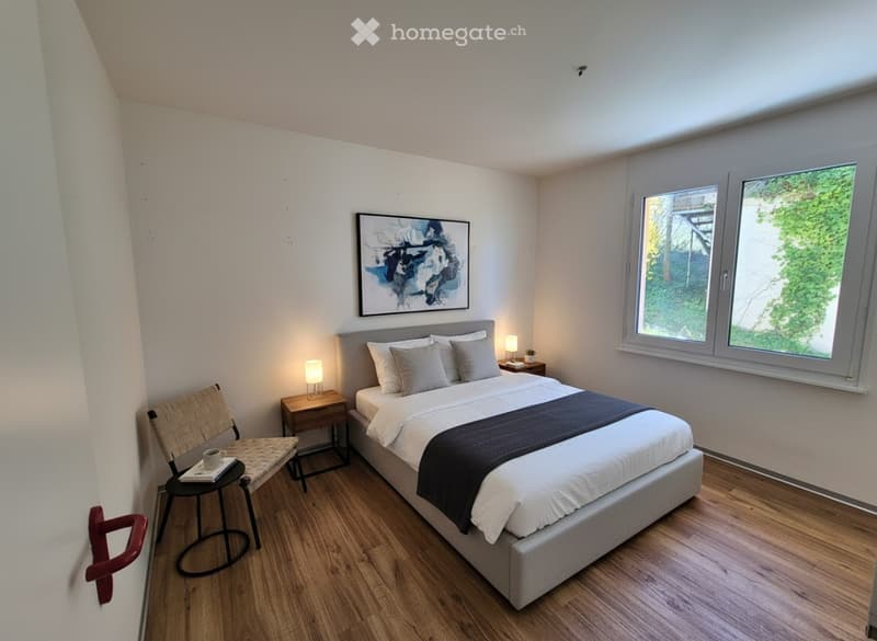 Modern bedroom with a large bed, nightstand, lamp, chair, and a small stool with a cup and books. The room features wooden flooring and large windows with a view of greenery.