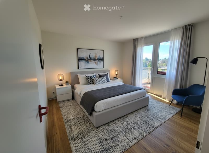Modern bedroom with a large bed, nightstand, armchair, floor lamp, and artwork on the wall. Large windows offer a view.