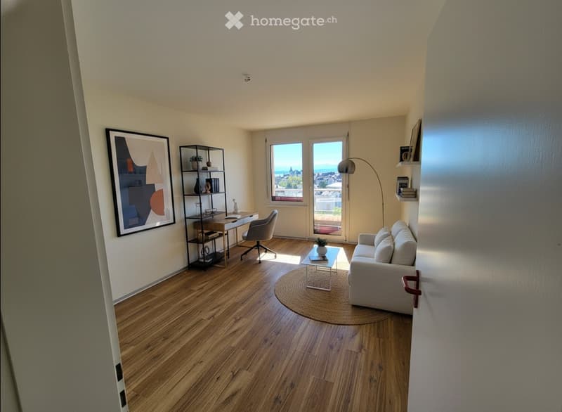 Bright living room with wooden flooring, large windows offering a scenic view, a sofa, coffee table, desk, and shelves.