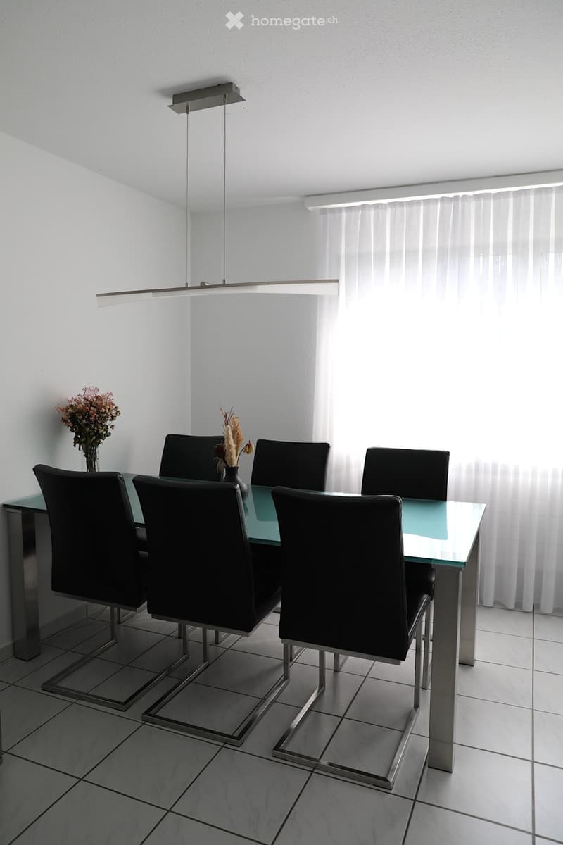 Dining room with a glass table, black chairs, white walls, white curtains, and a flower vase on the table
