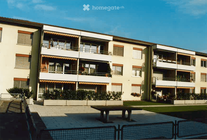 3 story apartment building, beige walls, multiple balconies, fenced garden area, table