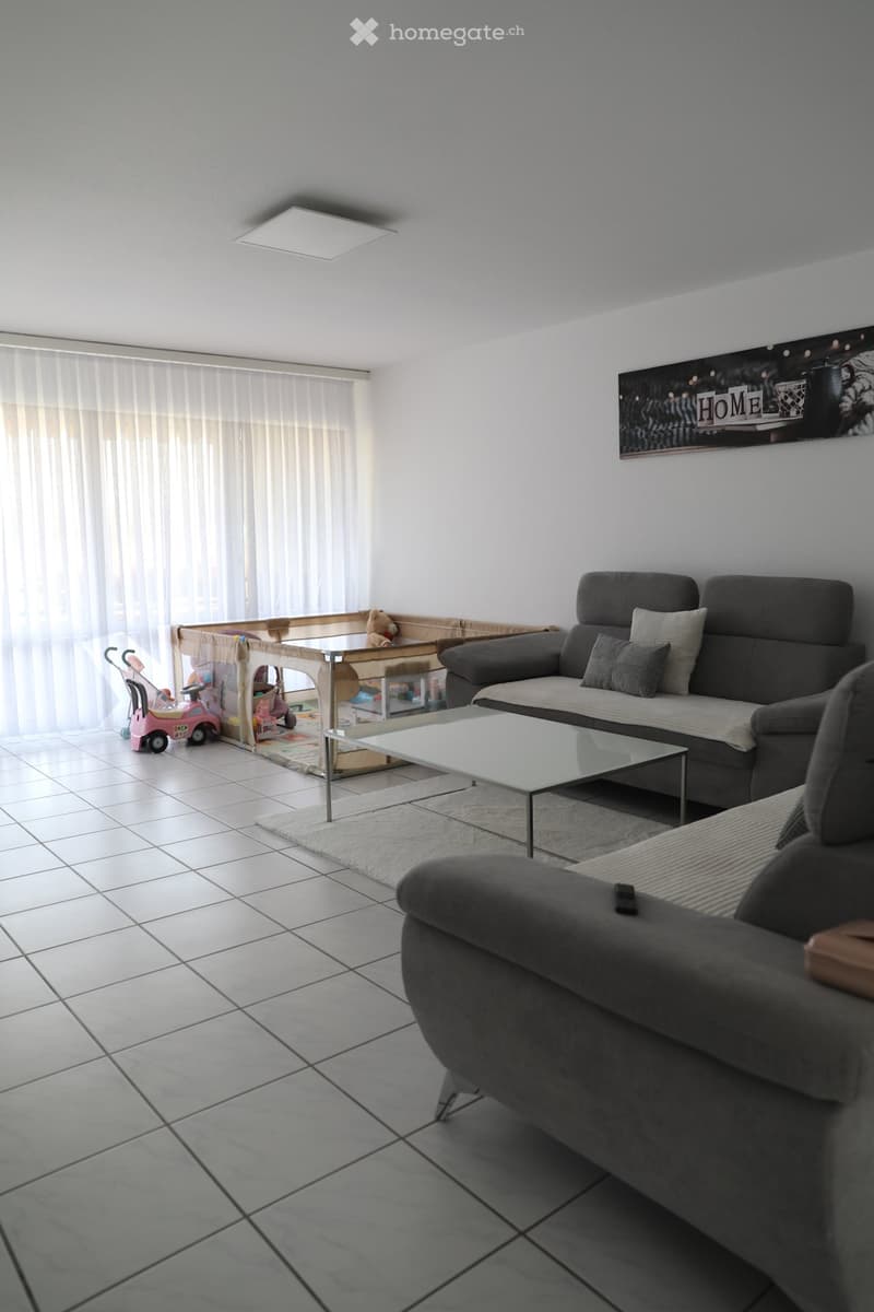 living room with white tiles, gray sofa, glass coffee table, white curtains, children's play area with toys