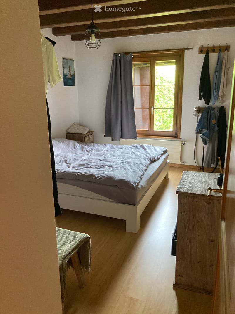 bedroom with bed, nightstand, wooden floor, window with curtains, wooden cabinet, clothes hanging on the wall