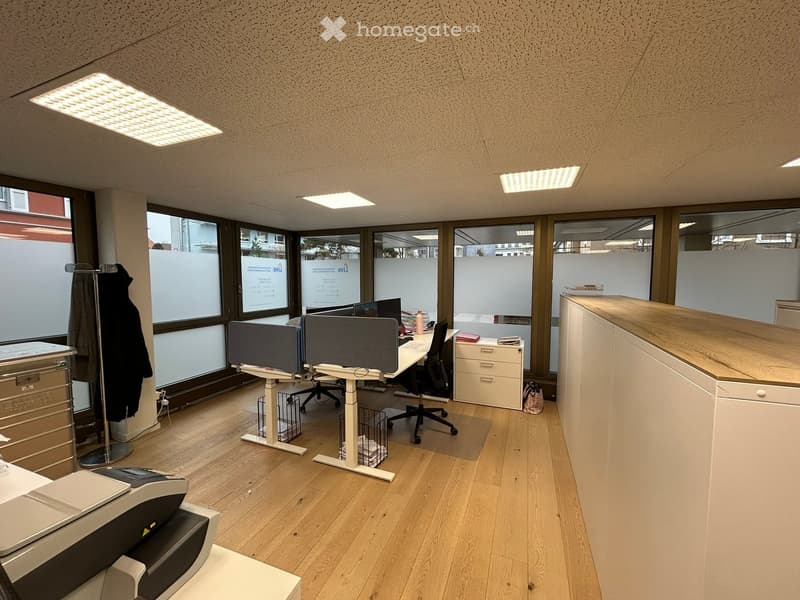 Office space with wooden floors, large windows, desks, chairs, cabinets, and a printer.