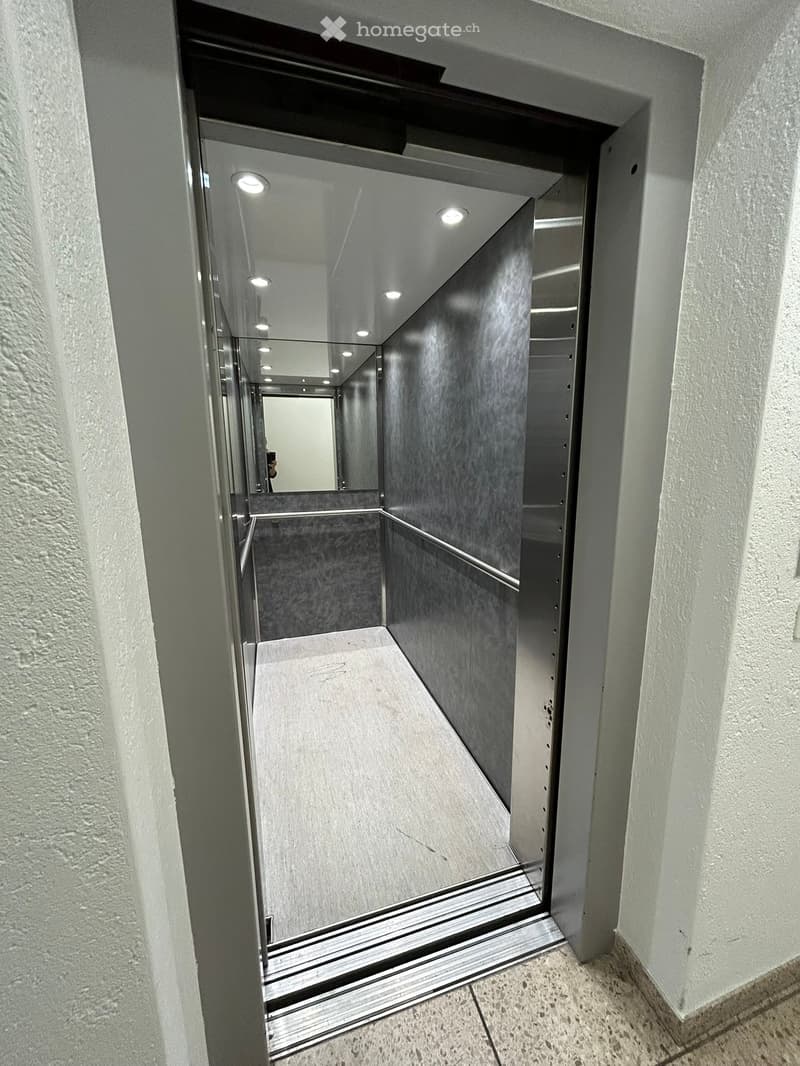 Stainless steel elevator with glass doors, white interior, and a mirror on the back wall.