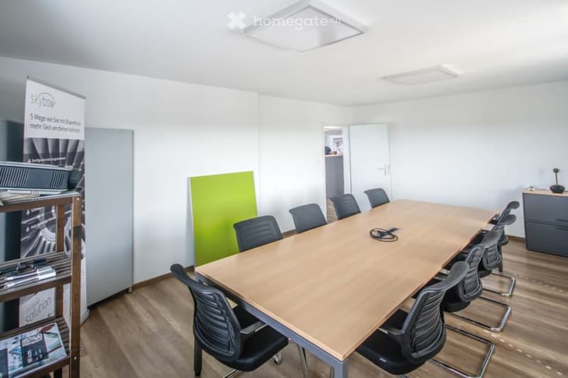conference room with a long table, chairs, wooden floor, white walls, ceiling lights, a green partition, a shelf with books, and a banner