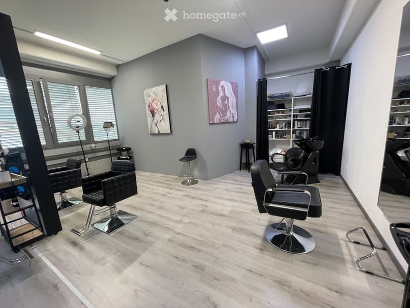 A salon with black leather chairs, a wooden floor, a gray wall, a shelf with bottles, and two paintings on the wall.